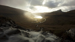 Mountain valley with rushing stream in foreground and glacial lake nestled between dark peaks under dramatic cloudy sky.