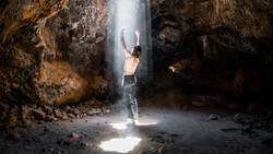 Shirtless man standing beneath a beam of sunlight in a cave, arms raised upward.