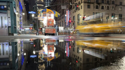 Wet city street at night with blurred yellow taxi and illuminated storefronts reflected in puddle.
