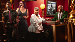 Four people in formal attire pose together in a warm-lit, burgundy-walled interior space decorated with gold accents.