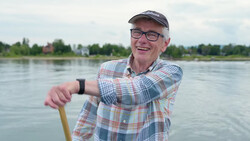 Smiling man in plaid shirt and cap holding paddle by river.