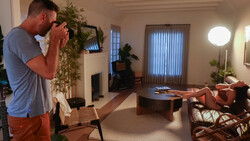 Photographer holding a camera with telephoto lens, composing a shot of a woman reclining on a couch in a modern living room.