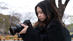 Female photographer holding a DSLR camera with telephoto lens, composing a shot outdoors.