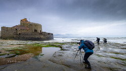 Photographer with tripod wading through tidal pools toward a stone fortress on a rocky coastline.
