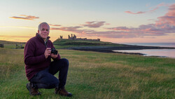 Photographer crouching in a grassy field at golden hour, holding a camera, with a ruined castle on a distant headland and water beyond.
