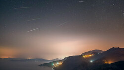 Meteor shower streaking across a twilight sky above a coastal landscape with illuminated hillside settlements.