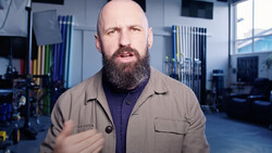 Bearded man in tan shirt speaking in a modern workshop studio with industrial shelving and blue-tinted lighting.
