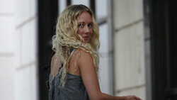 Woman with long blonde wavy hair wearing a gray sleeveless top, looking over her shoulder in an indoor hallway.