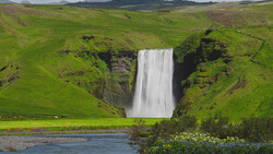 Powerful waterfall cascading into a river surrounded by vivid green hillsides and dark basalt cliffs.