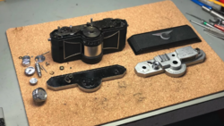 Disassembled rangefinder camera components laid out on cork surface showing film chamber, lens barrel, viewfinder, and metal mechanical parts.