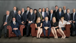Professional group portrait of approximately 20 business professionals in formal attire arranged in three rows.
