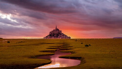 Medieval hilltop castle silhouetted against dramatic sunset sky with winding stream in foreground.