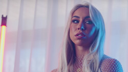 Woman with long platinum blonde hair looking away against neon-lit backdrop with vertical blinds.