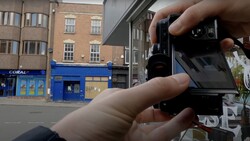Hands holding a vintage film camera with the back open, showing the film compartment against a street scene with brick buildings and a blue storefront.