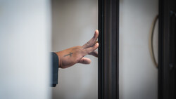 Hand reaching through a dark doorframe against white walls, fingers extended in an open gesture.