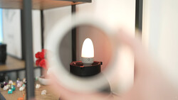 LED candle with glowing white flame inside dark holder, viewed through blurred circular glass or lens.