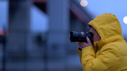 Photographer in yellow rain jacket holding camera to eye during twilight urban shoot.