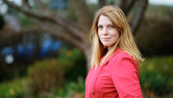 Woman with strawberry blonde hair wearing a bright pink blazer, photographed outdoors against a blurred garden background.