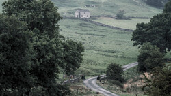 Winding road through rolling green hills with stone house and trees framing the valley.
