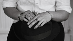 Close-up of two people's hands with wedding rings resting on a dark hat, shot in black and white.
