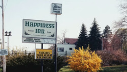 Vintage motel sign for Happiness Inn with yellow flowering shrub and evergreen trees in background.