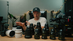 Filmmaker demonstrating cinema lenses arranged on wooden desk in studio setting.