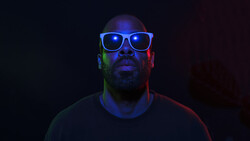 Man wearing illuminated blue-framed glasses against dark background with colored gel lighting.
