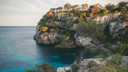 Limestone cliff with turquoise cove and Mediterranean vegetation overlooking calm blue sea.