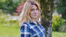 Woman in blue plaid shirt leaning against a moss-covered tree trunk in a garden setting.