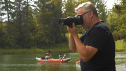 Photographer with telephoto lens capturing kayakers on a river surrounded by evergreen trees.