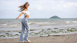 Woman in white off-shoulder top and jeans running on beach with ocean and distant peninsula in background.