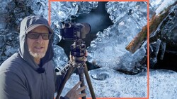 Photographer in winter gear holding a camera on a tripod amid ice-covered rocks and frozen surfaces.