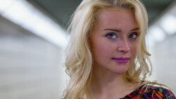 Blonde woman with purple-toned makeup looking directly at camera in soft studio lighting.