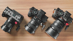 Three cinema cameras with large telephoto lenses arranged on a wooden surface, each equipped with top-mounted monitoring screens and red lens caps.