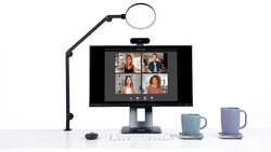 Video conferencing setup with monitor displaying four participants, ring light on articulating arm, keyboard, and two mugs.