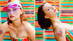 Two women posed against a vibrant multicolored striped background, wearing bold makeup and shoulder tattoos.