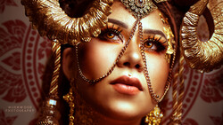 Woman wearing elaborate gold headdress and ornate jewelry with dramatic eye makeup against patterned backdrop.