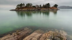 Rocky foreground with small forested island and its reflection in calm green water under overcast sky.