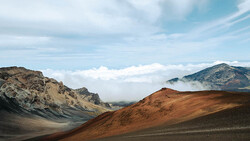 Volcanic landscape with layered rust and charcoal-colored cinder cones beneath a cloud-filled sky.