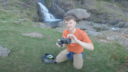 Photographer kneeling outdoors holding a camera, with a small blue camera bag beside him and a waterfall visible in the background.