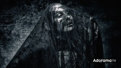 Figure draped in tattered black fabric against dark textured background with dramatic side lighting.