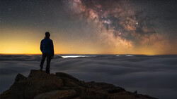 Silhouetted figure standing on rocky outcrop beneath the Milky Way galaxy at night.