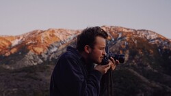 Photographer holding a telephoto lens to his eye, positioned against a mountainous desert landscape at golden hour.