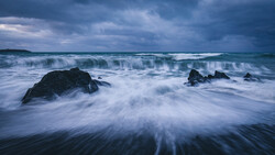 Waves crash over dark rocks on a beach under a dramatic stormy sky.