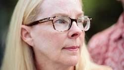 Woman wearing eyeglasses with blonde hair, looking thoughtfully to the side against a blurred green background.