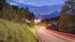 Illuminated church with spire surrounded by forested hills and a winding road with light trails at dusk.
