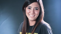Young woman smiling at camera wearing dark graphic t-shirt against blue background.