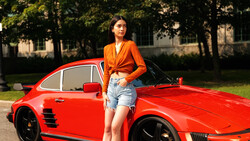 Woman in orange shirt leaning against bright red sports car in outdoor setting.
