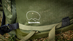Close-up of a green Shimoda camera bag showing the brand logo and webbing straps with black plastic hardware.