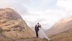Bride and groom embracing in a mountain valley between two steep peaks under partly cloudy sky.